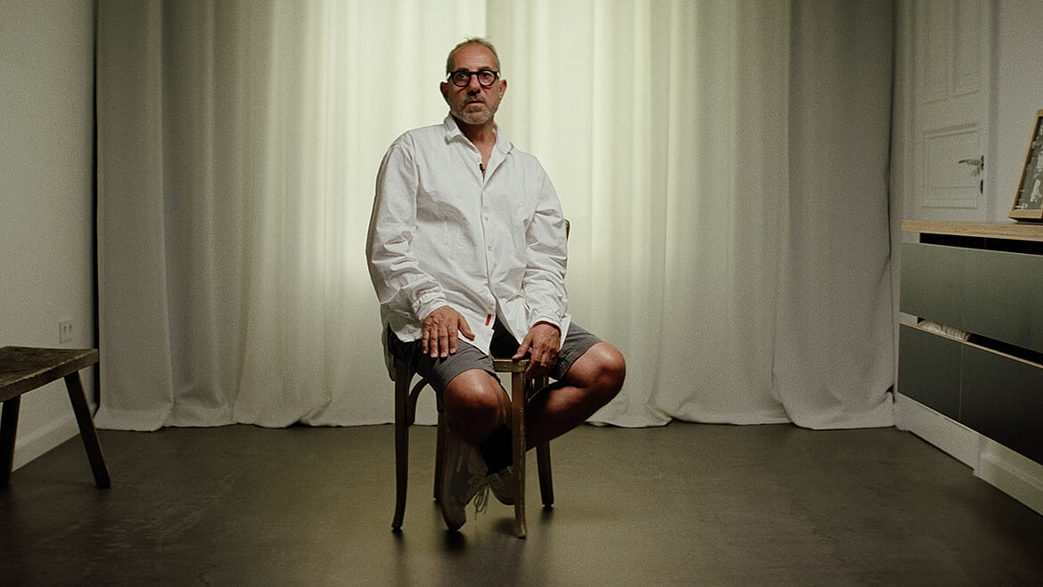 Person seated on a chair in a minimal interior space with a light-colored wall, facing the camera in a calm atmosphere