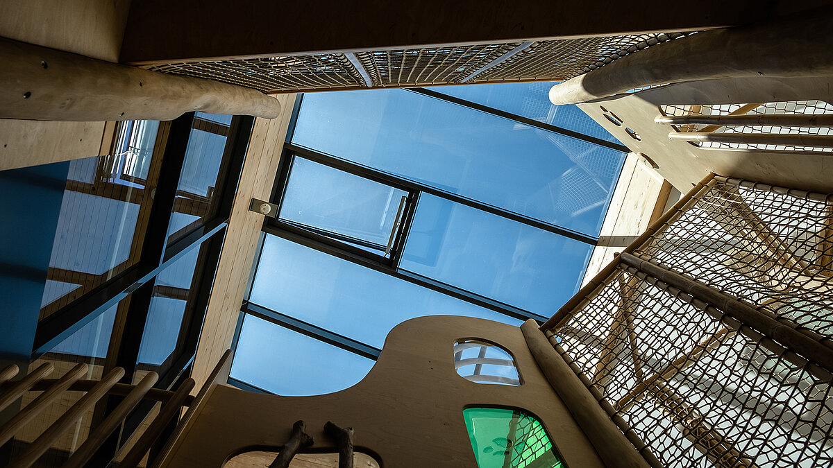 Kita Emilio with large glass roof and integrated ventilation opening above a wooden play structure in a daylight-filled interior