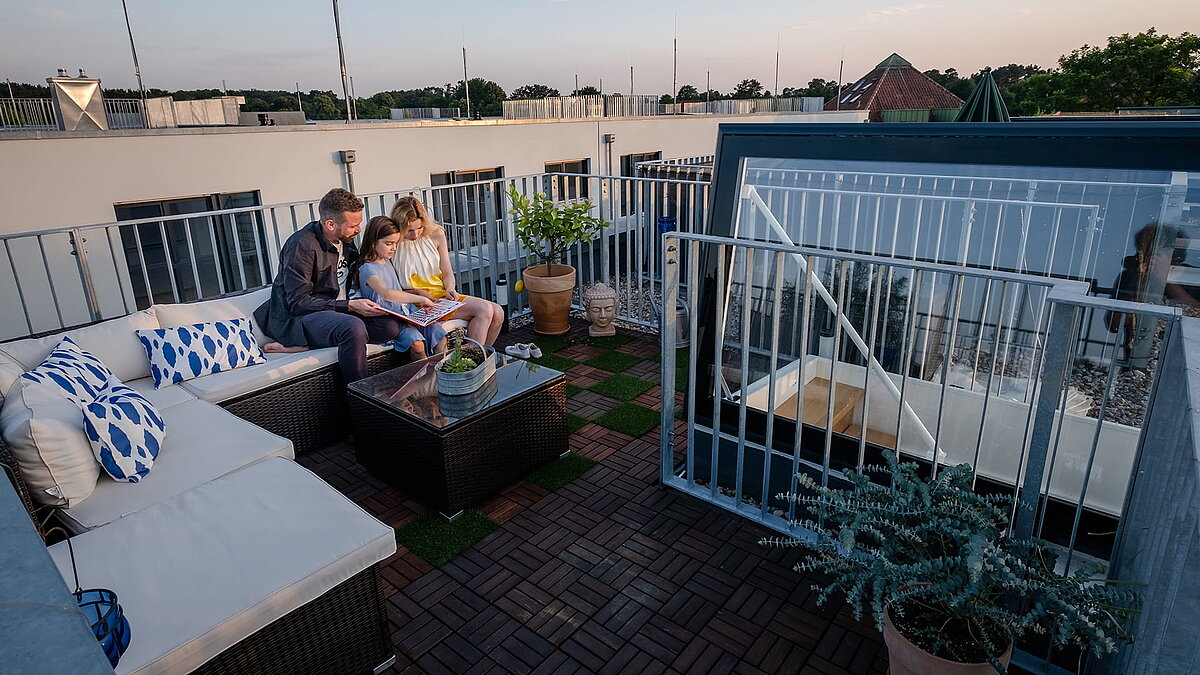 A family sits on the cosy roof terrace with seating area of their townhouse in Berlin with flat roof exit at sunset.