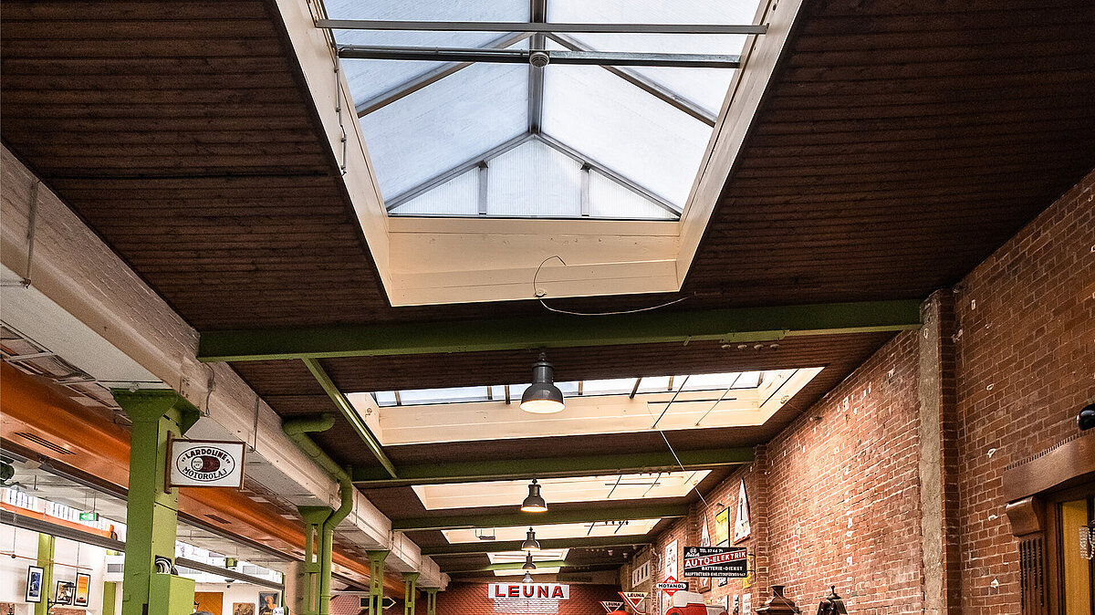 Interior view of the Zündmagnet event hall in Wurzen featuring a glass roof, green steel beams, and brick walls with industrial charm.