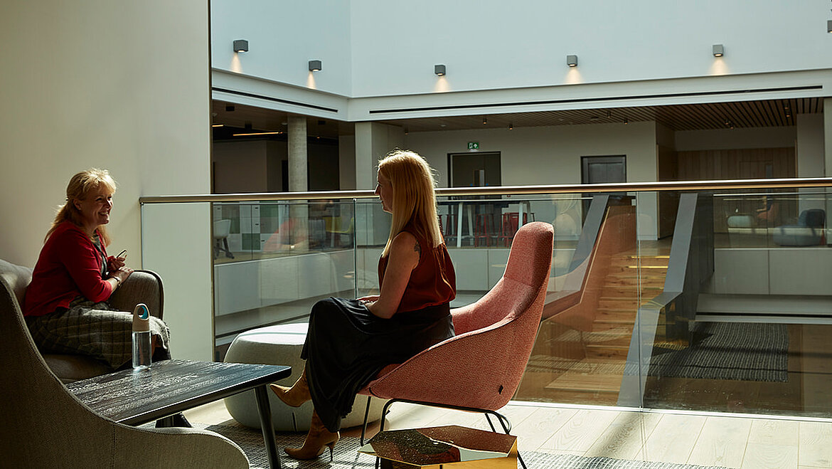Two women converse in a modern, light-filled lounge with skylights