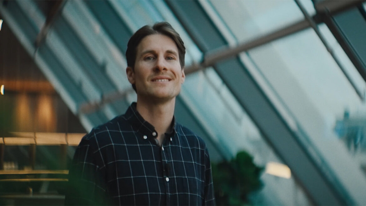Person standing inside a modern building with a glass roof, looking slightly upward with a subtle smile as natural daylight enters through the roof glazing