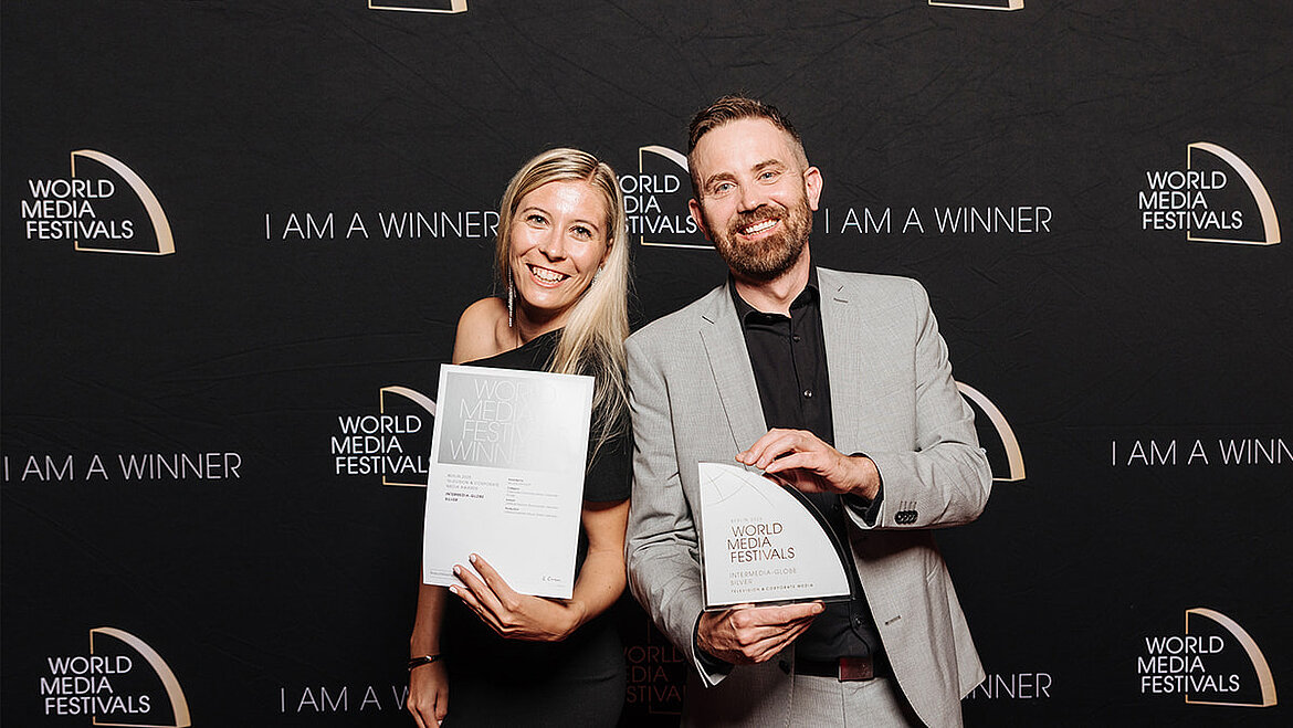 Two people standing side by side in front of a dark event backdrop, holding an award and a framed certificate