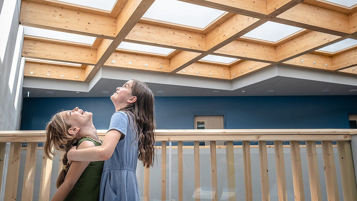 Two laughing girls under a modern wooden skylight ceiling with natural daylight