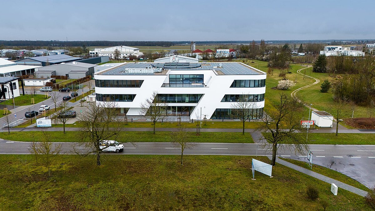 Modern office building with white facade and solar system on the roof, surrounded by green spaces and streets, under a cloudy sky.