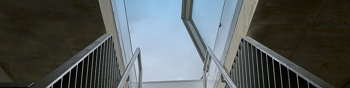 Upward view of a concrete staircase with metal railings leading to an open LAMILUX flat roof access hatch.