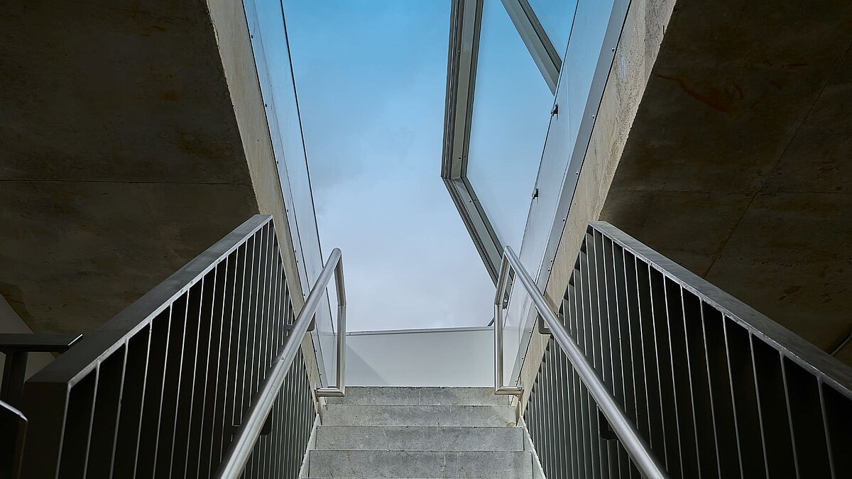 Upward view of a concrete staircase with metal railings leading to an open LAMILUX flat roof access hatch.