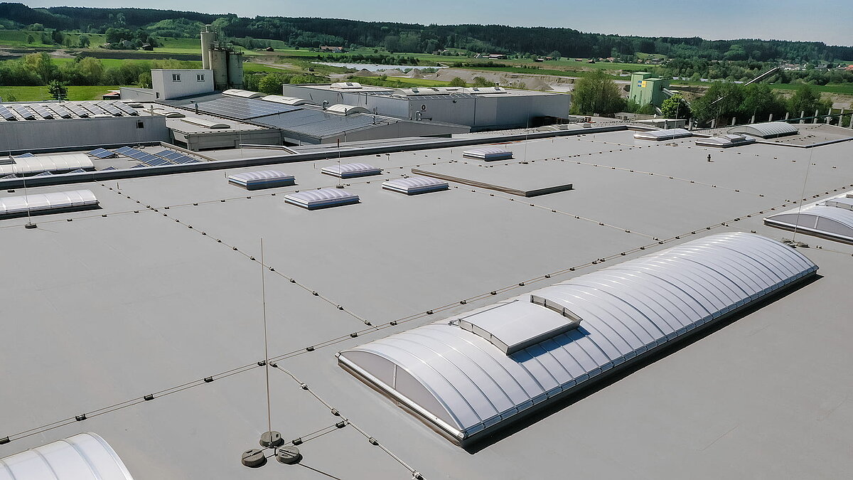 Roof of the Unglehrt construction company's production hall featuring LAMILUX F100 W skylights and B continuous rooflights, surrounded by a green landscape.