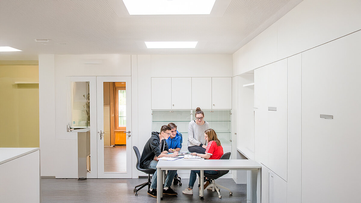 Bright interior view of the Markgraf-Georg-Friedrich-Gymnasium in Kulmbach with students working at a table under flat roof windows.