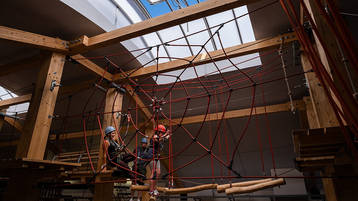 Interior view of the "Alte Brauerei" climbing hall in Eisenach featuring a LAMILUX skylight band B and three people on a high ropes course with a climbing net.