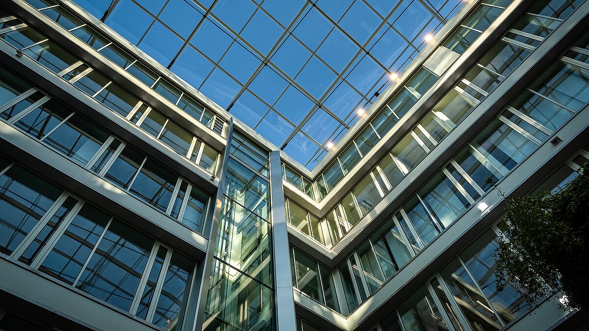 Upward view of the modern glass roof and facades of the Newton office building of the TÜV Süd Group in Munich, featuring reflective windows and a blue sky.