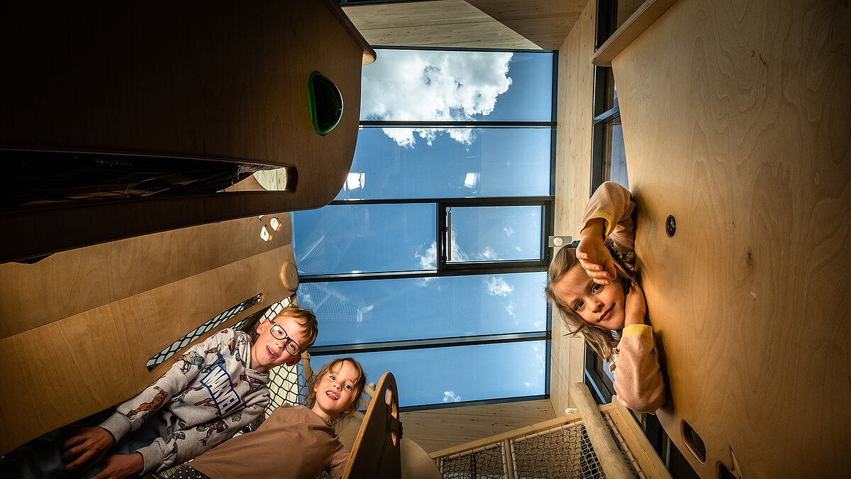 Children looking down into the camera, above them a large glass roof with a view of the sky, bright interior with wooden finishes