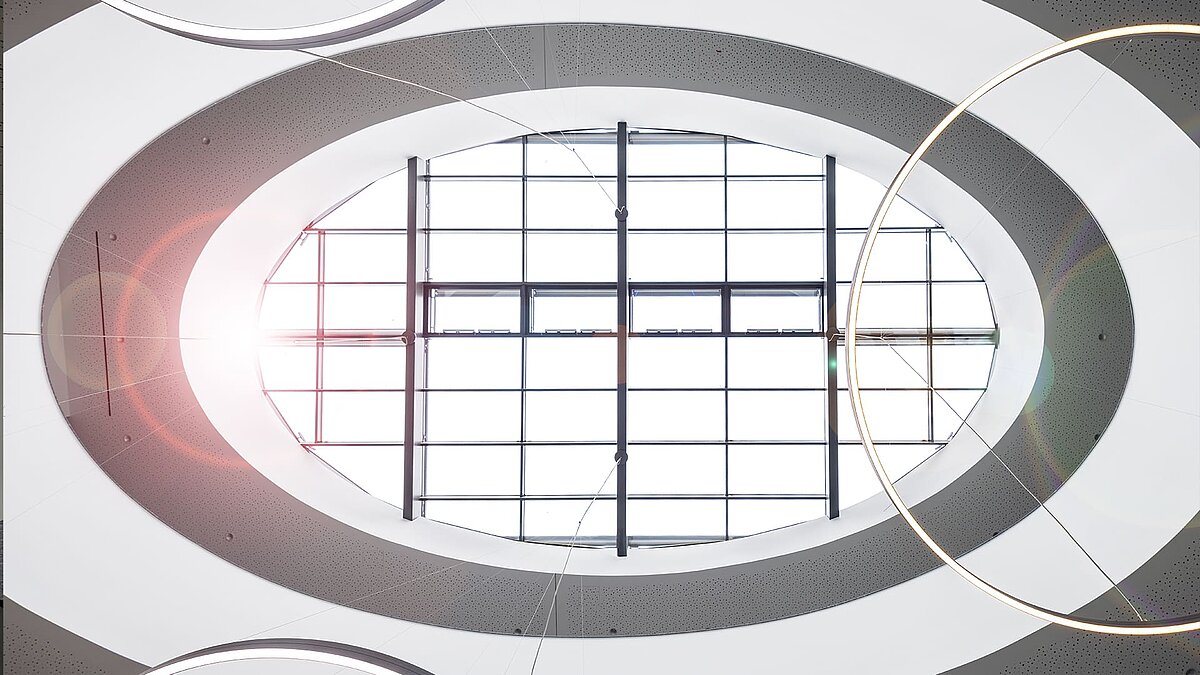 [Translate to English:] Blick nach oben auf ovales Glasdach mit Sonnenlicht und modernen Ringlampen im Atrium eines Bürogebäudes.