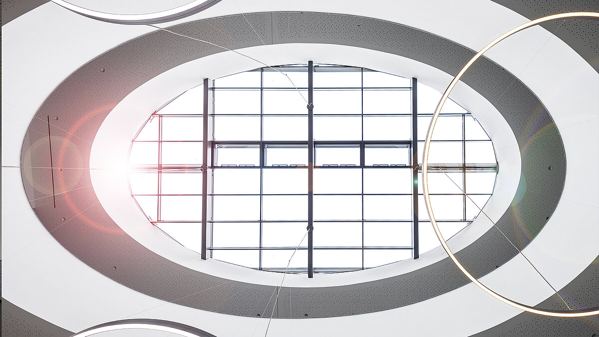 [Translate to English:] Blick nach oben auf ovales Glasdach mit Sonnenlicht und modernen Ringlampen im Atrium eines Bürogebäudes.