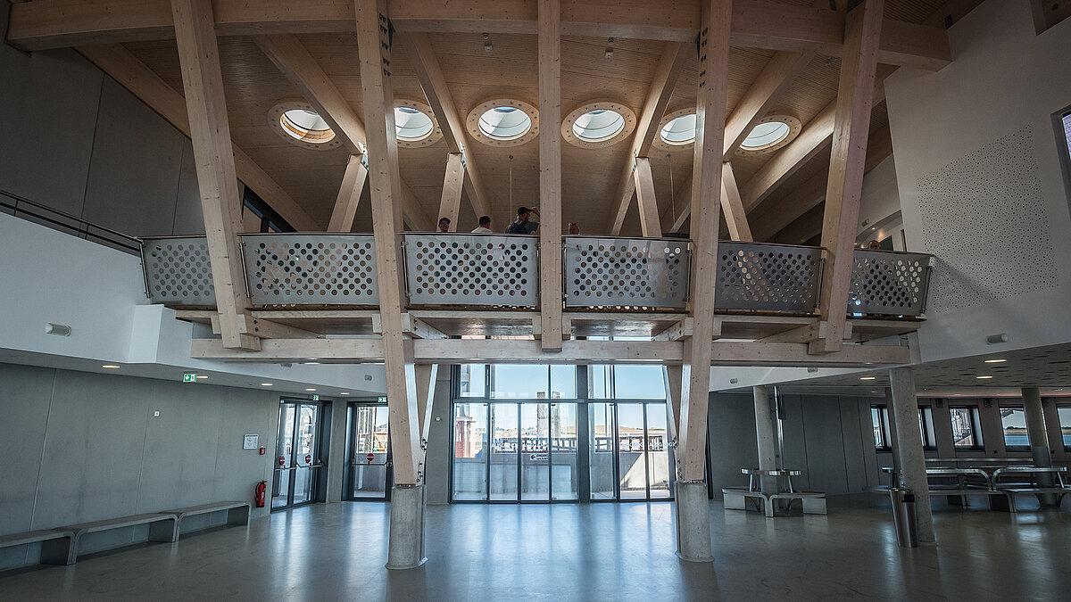 Interior of the Norderney harbour terminal with visible wooden beams, round flat roof windows and modern railings.