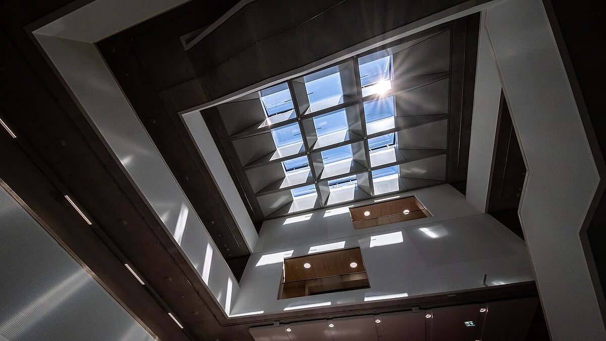 Interior view of a modern atrium with glass roof, wooden beams, and exposed concrete walls