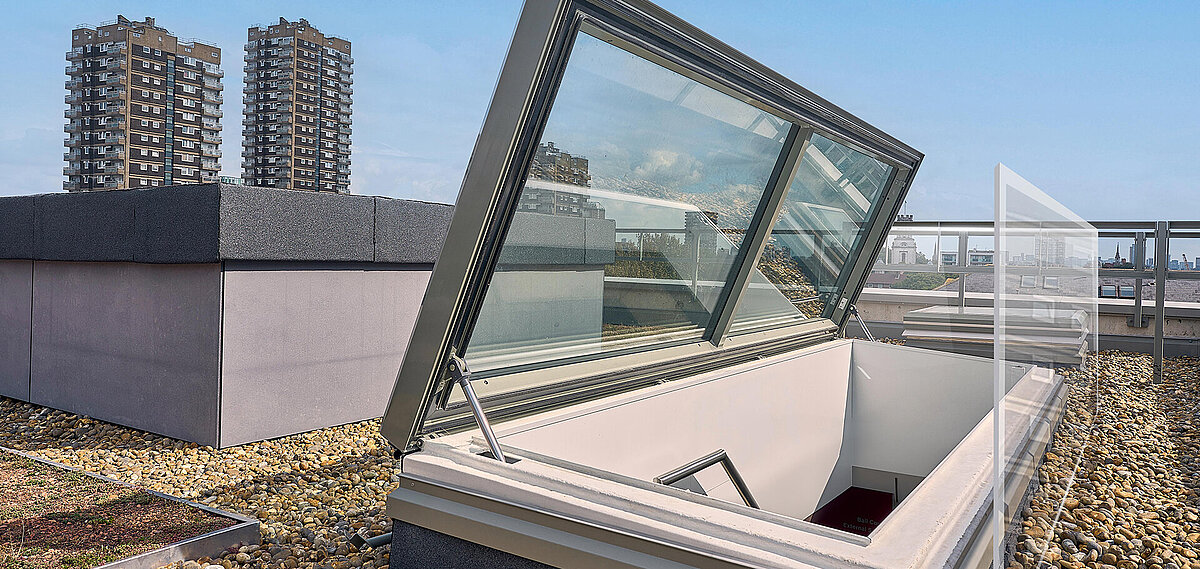 Open LAMILUX flat roof access on a green and gravel-covered rooftop, with high-rise buildings and the London skyline in the background.