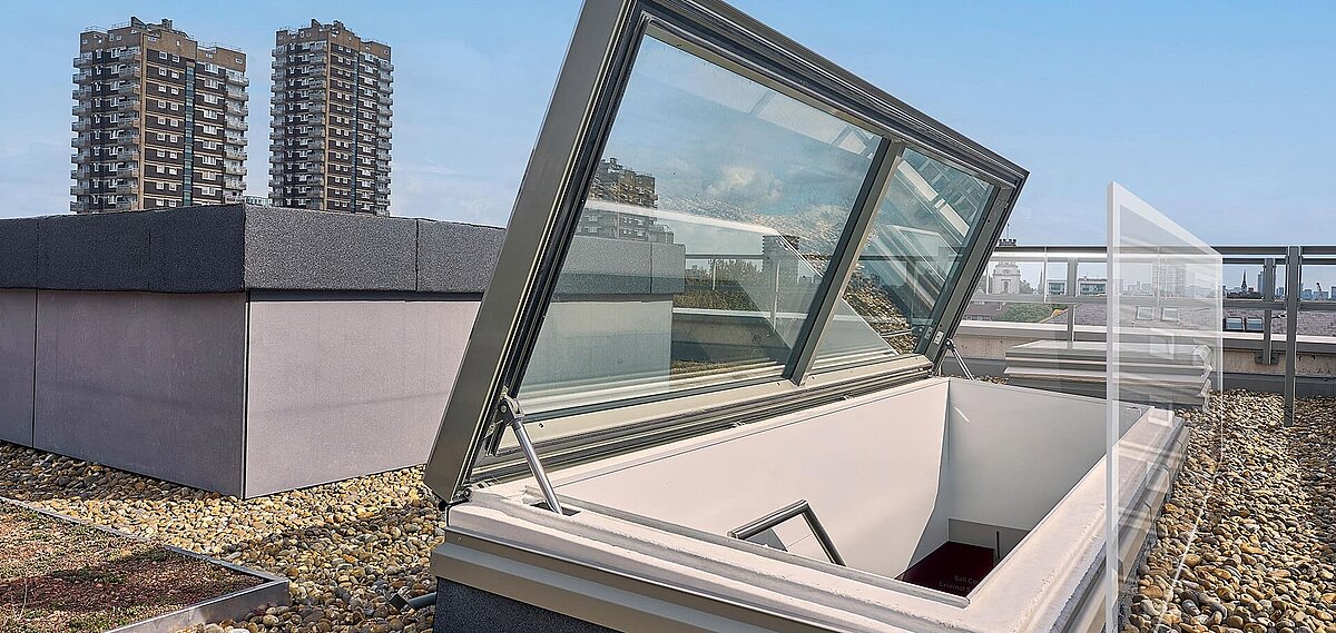 Open LAMILUX flat roof access on a green and gravel-covered rooftop, with high-rise buildings and the London skyline in the background.