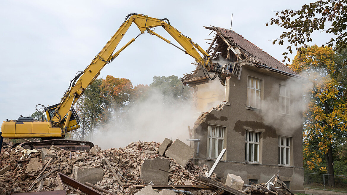 An excavator stands on a pile of rubble and demolishes a grey building.