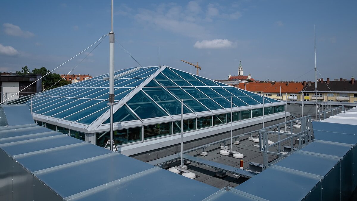  Glass roof of the Dante-Gymnasium with a stable steel structure and blue glazing, surrounded by buildings with a blue sky in the background.