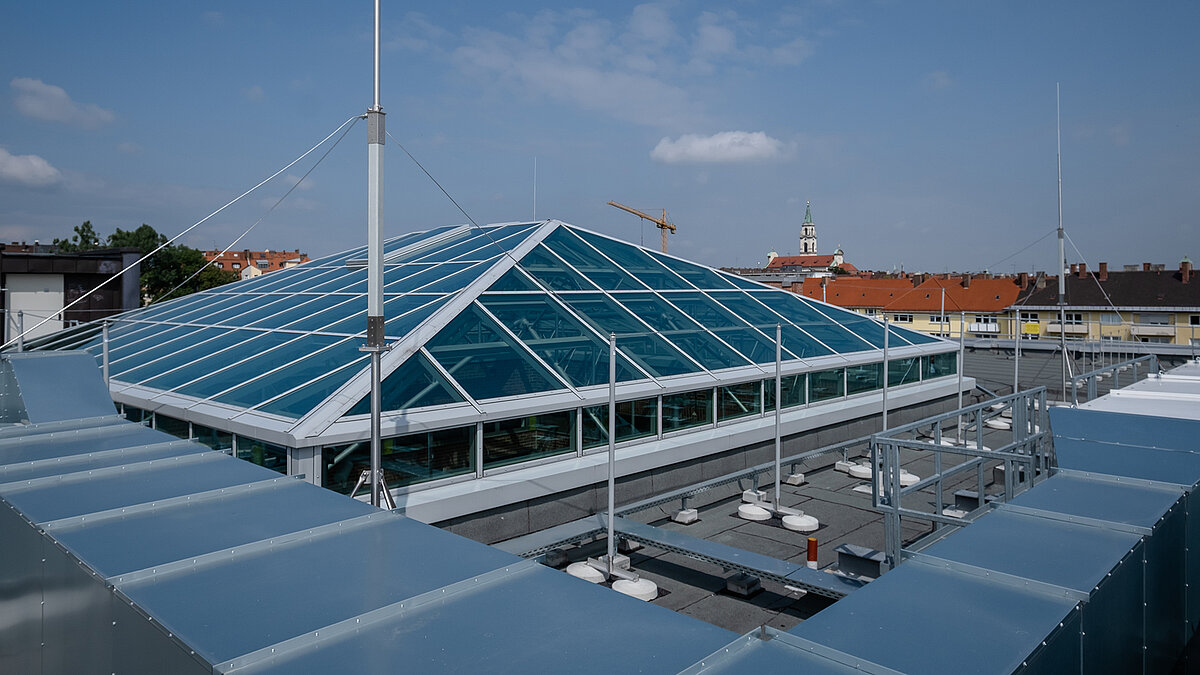  Glass roof of the Dante-Gymnasium with a stable steel structure and blue glazing, surrounded by buildings with a blue sky in the background.