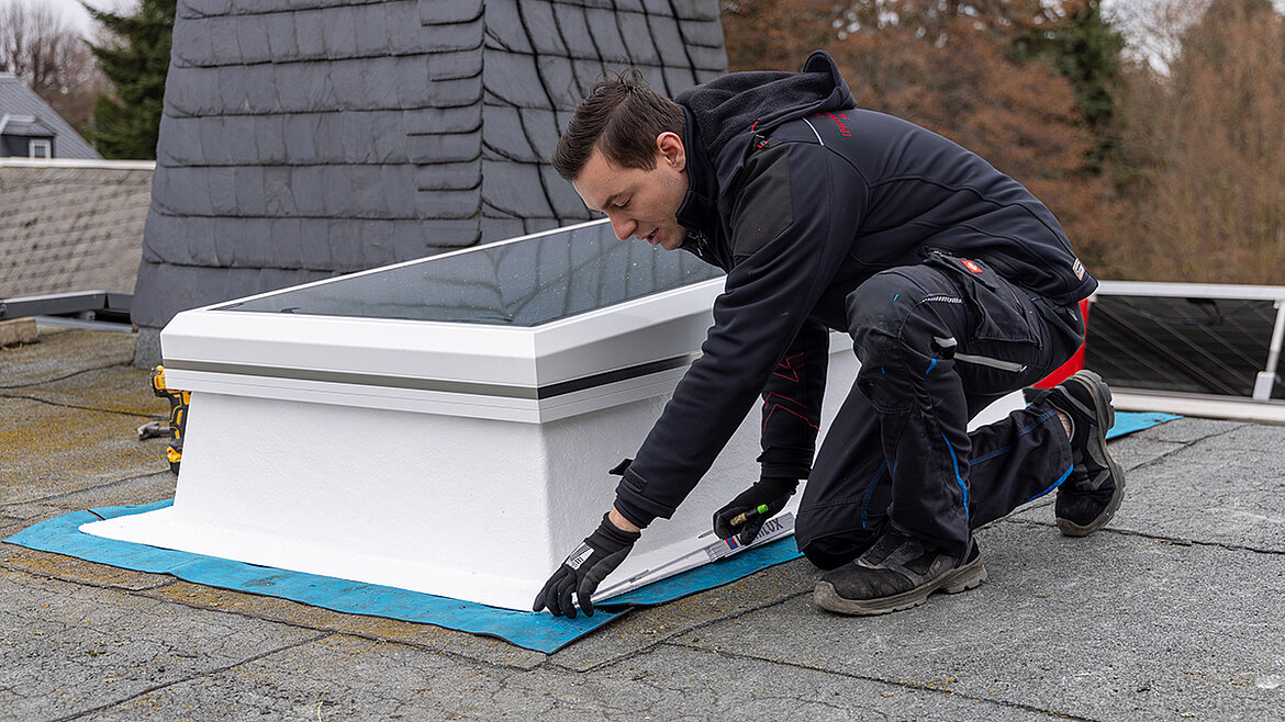 A craftsman kneels on a flat roof and installs a skylight
