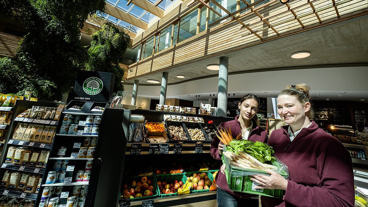 Two staff members at the Byodo organic food store in Mühldorf, showcasing fresh organic vegetables in a modern, light-filled shopping area with wood and glass accents.