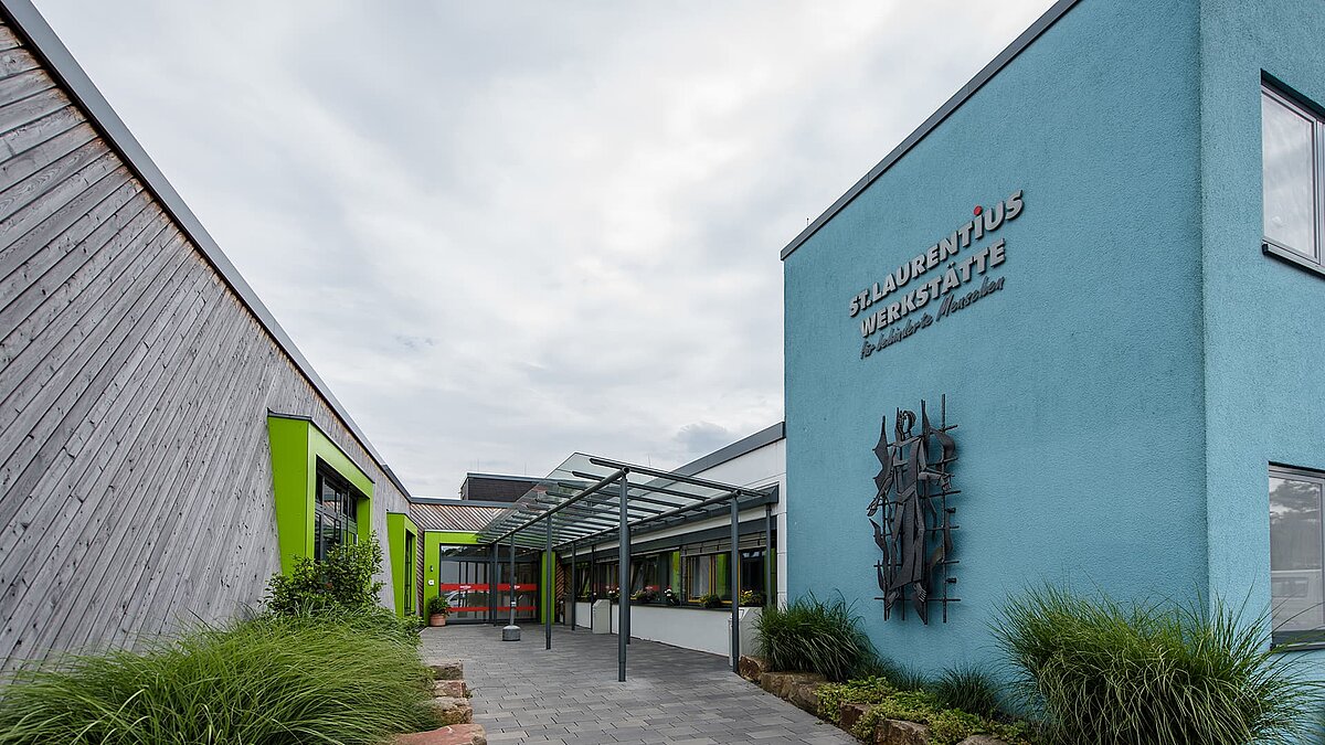 Entrance area of the St Laurentius workshop of the Caritas association in Hagen with glass canopy, blue façade and green window frames, surrounded by plants.