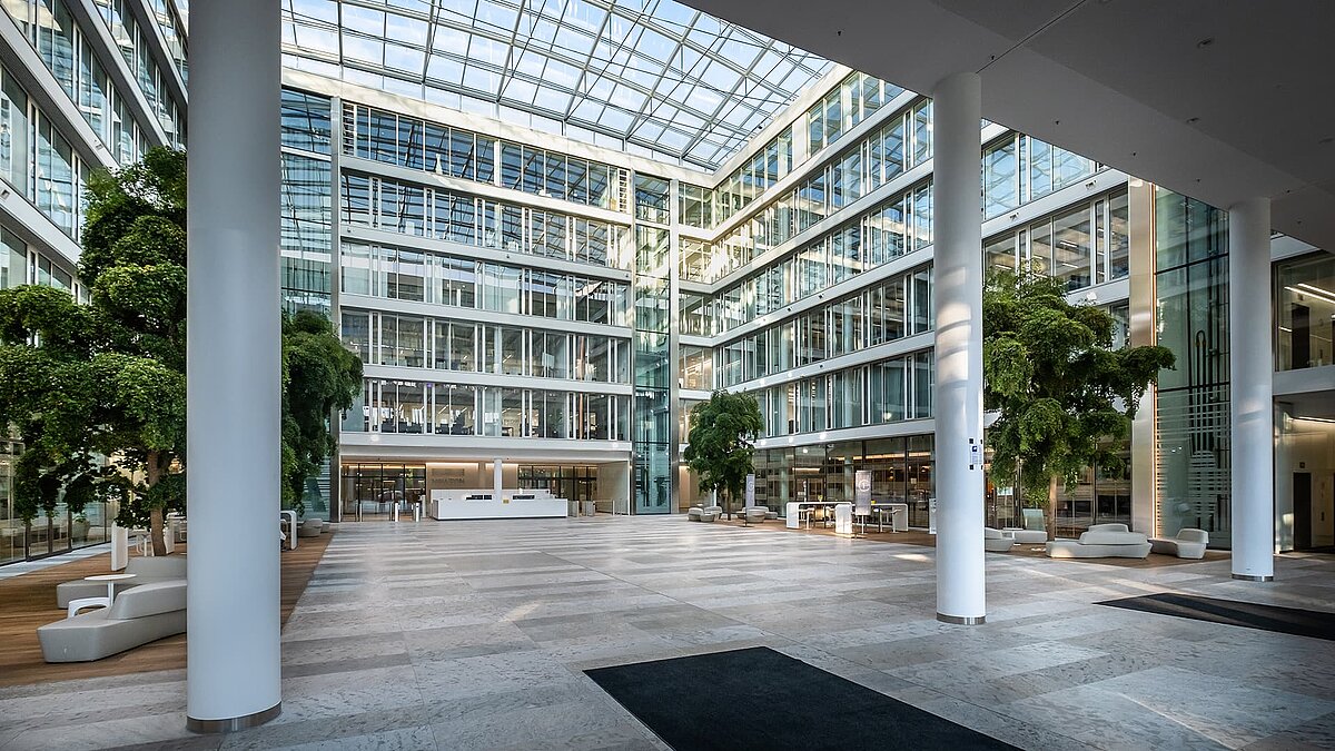 Large atrium in the Newton office building of the TÜV Süd Group in Munich with a glass roof, modern columns, and green interior spaces.