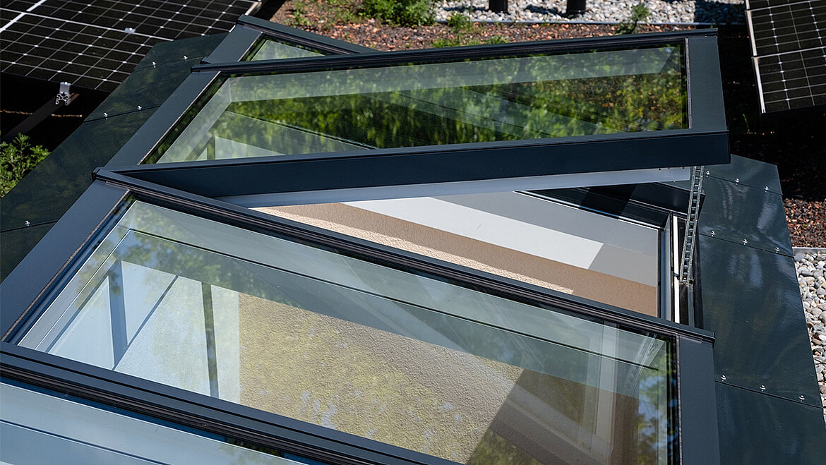 An open aluminium skylight strip on a flat roof with solar panels, a gravel bed and extensive green roofing in the background