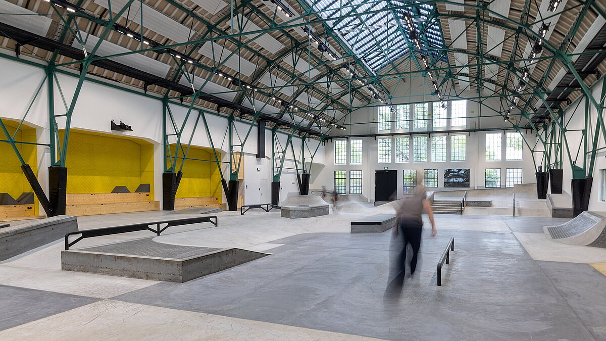 Skate hall inside renovated industrial building with glass roof and green steel structure.
