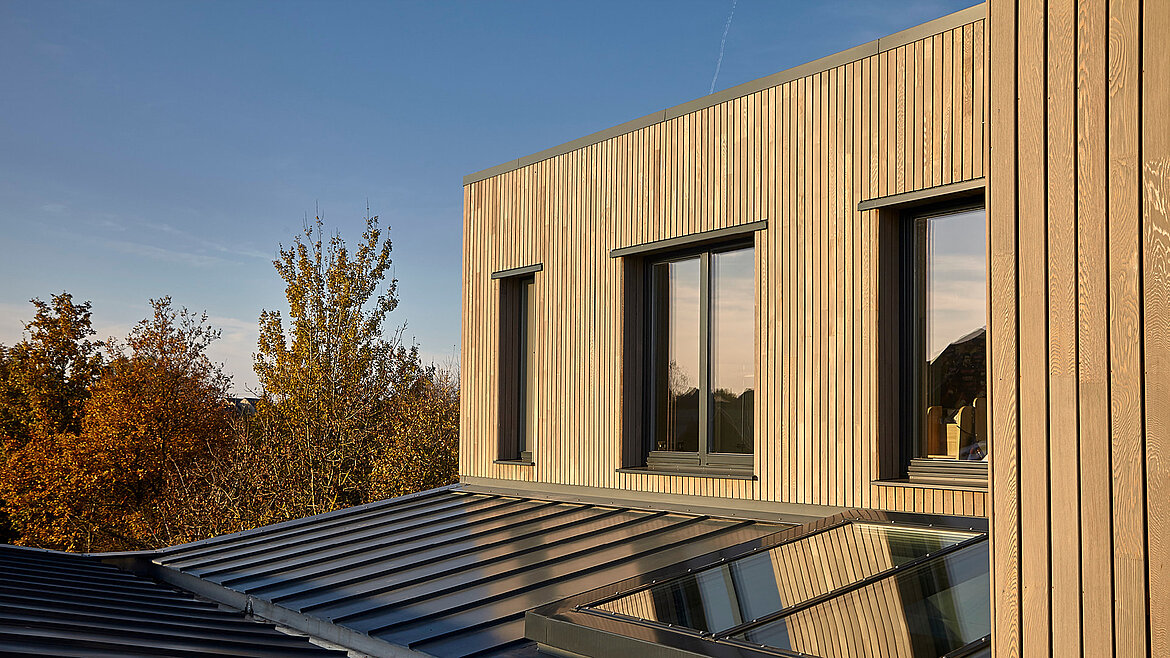 Modern wooden facade with large windows and glass roof in the foreground at sunset.