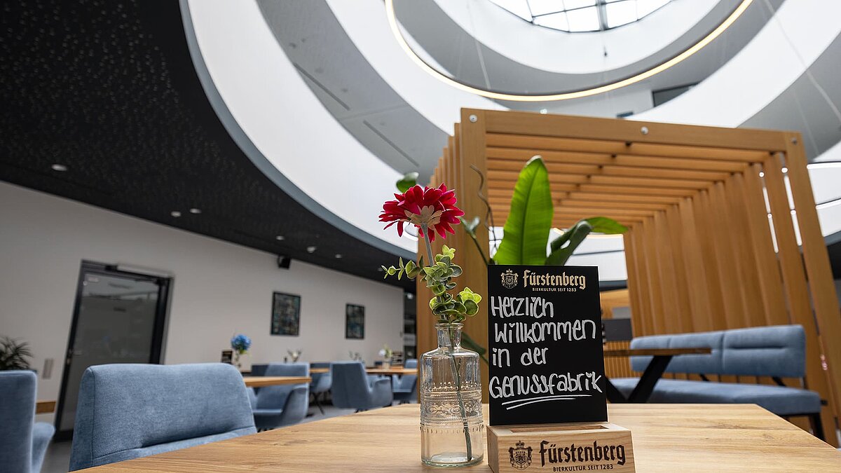 Interior view of a modern dining area with circular skylights, wooden elements, and a sign reading "Welcome to the Genussfabrik".