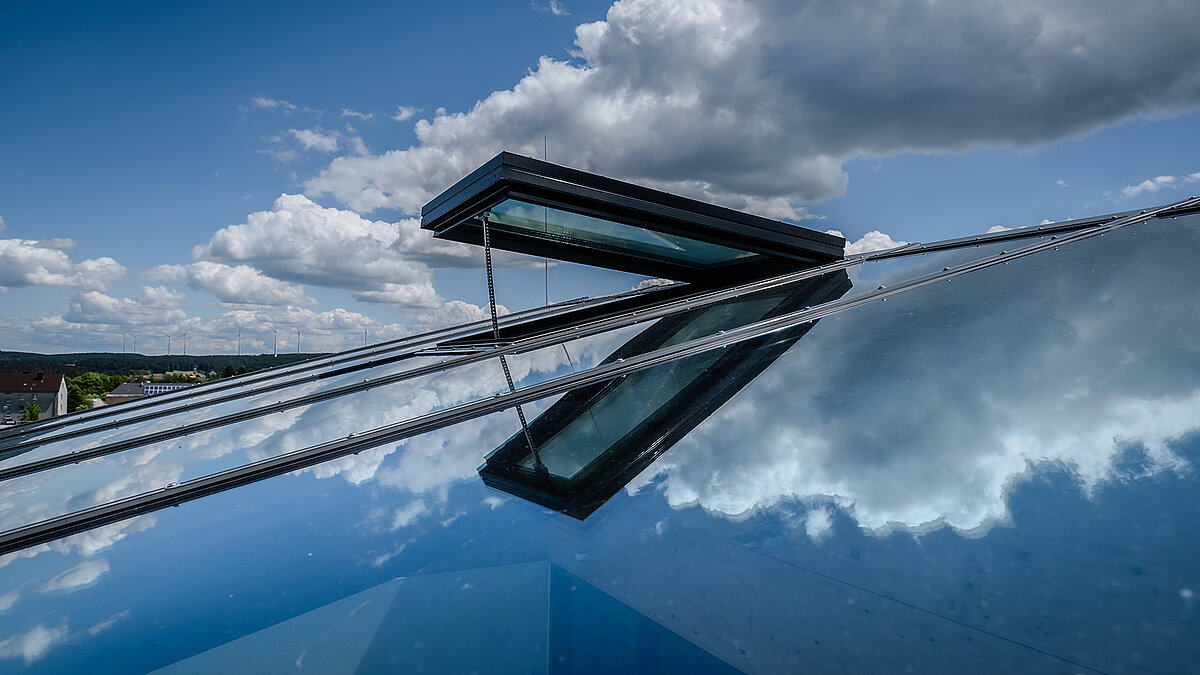 Close-up of a sloped glass roof with an open ventilation flap with clouds reflected in the glazing