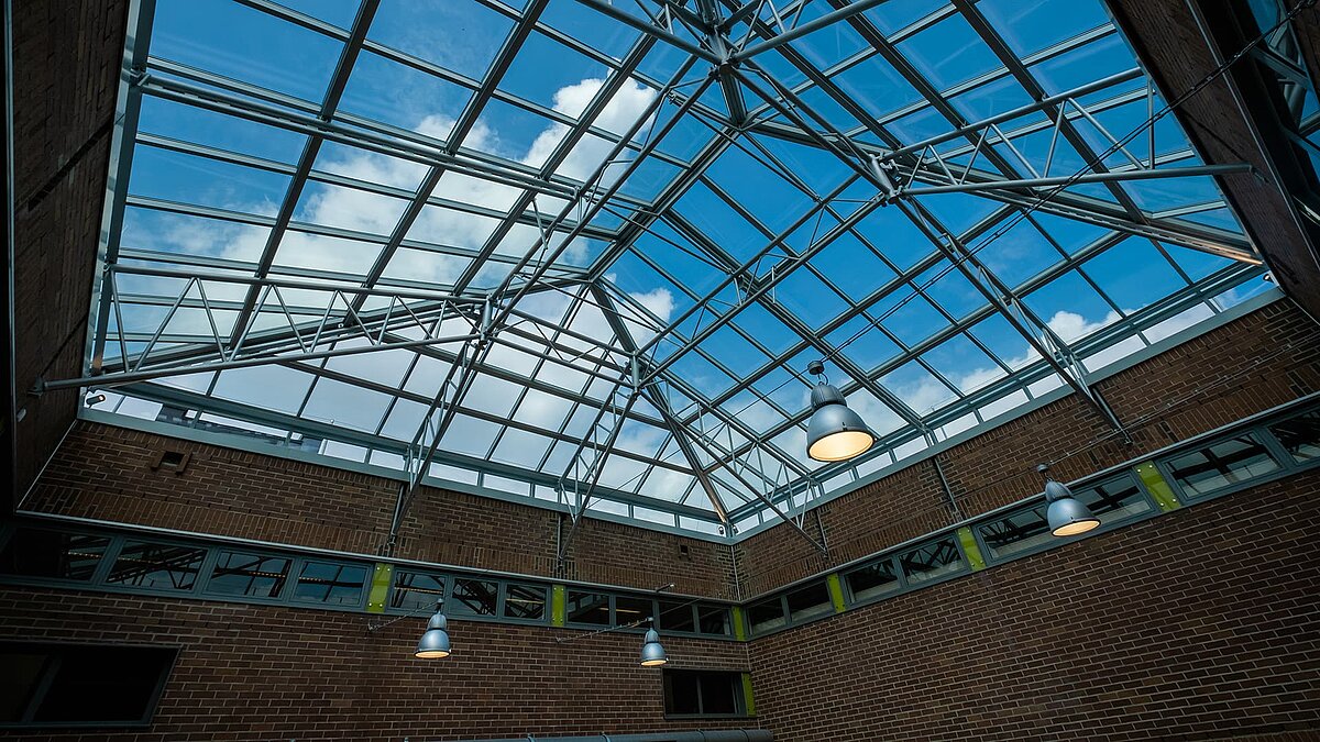 Glass roof of Dante-Gymnasium with steel beams and modern pendant lights, view of blue sky.