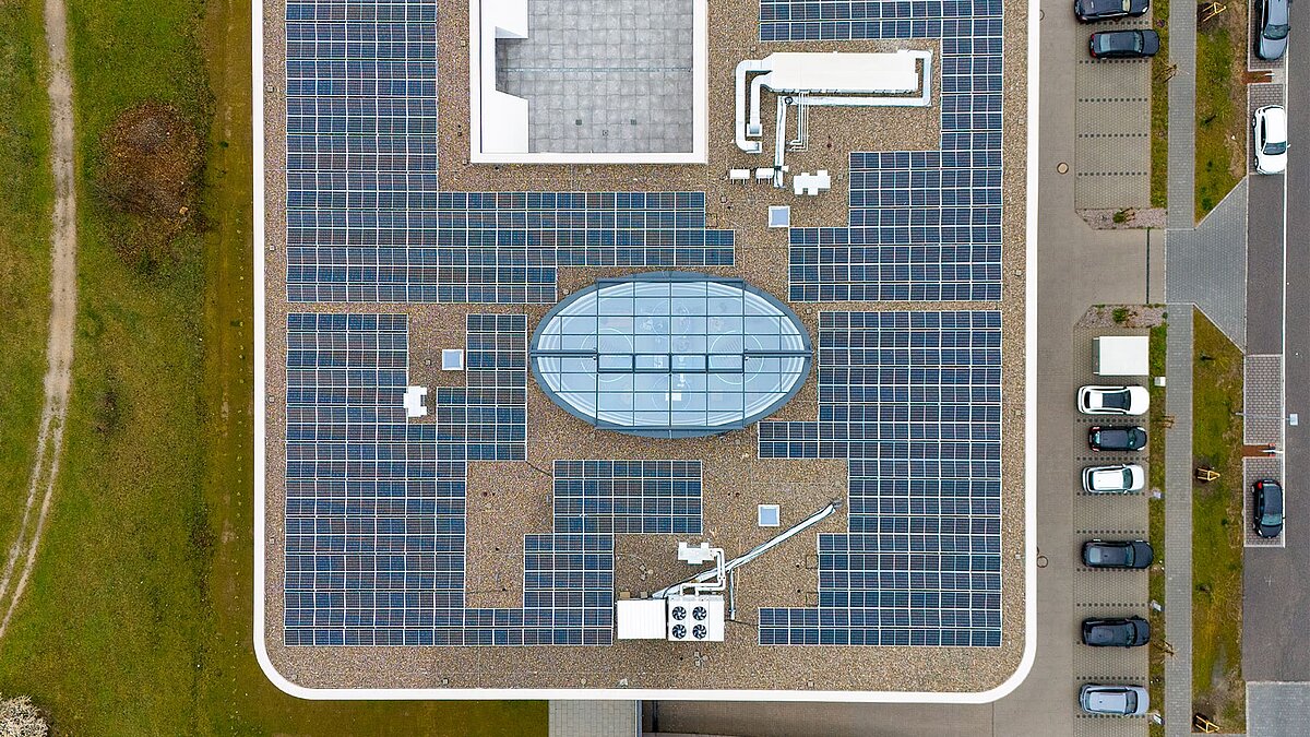 Drone view of a flat roof with numerous solar panels and an oval glass structure in the center, with a parking lot and cars next to it.