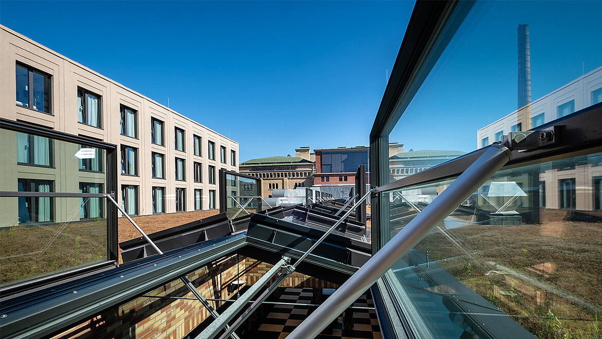 An open glass roof with a metal frame, looking out over a green flat roof and modern office buildings beneath a blue sky