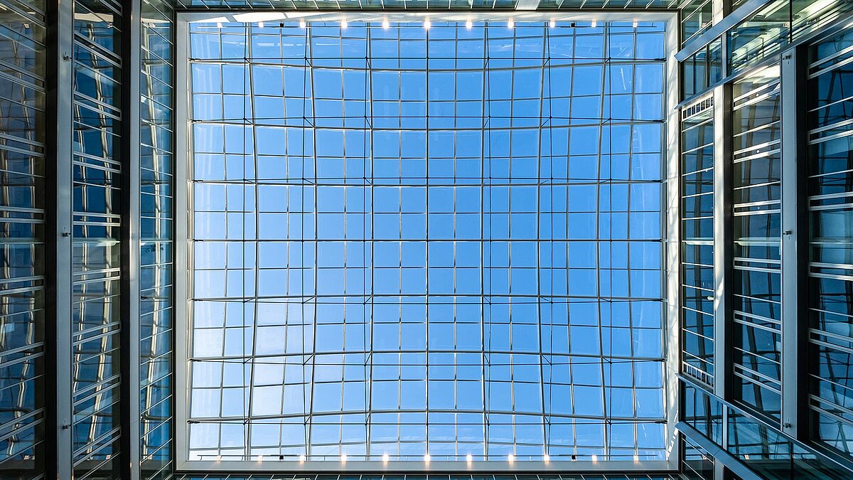 Upward view of the glass roof in the atrium of the Newton office building of the TÜV Süd Group in Munich, featuring a blue sky and modern glass structures.