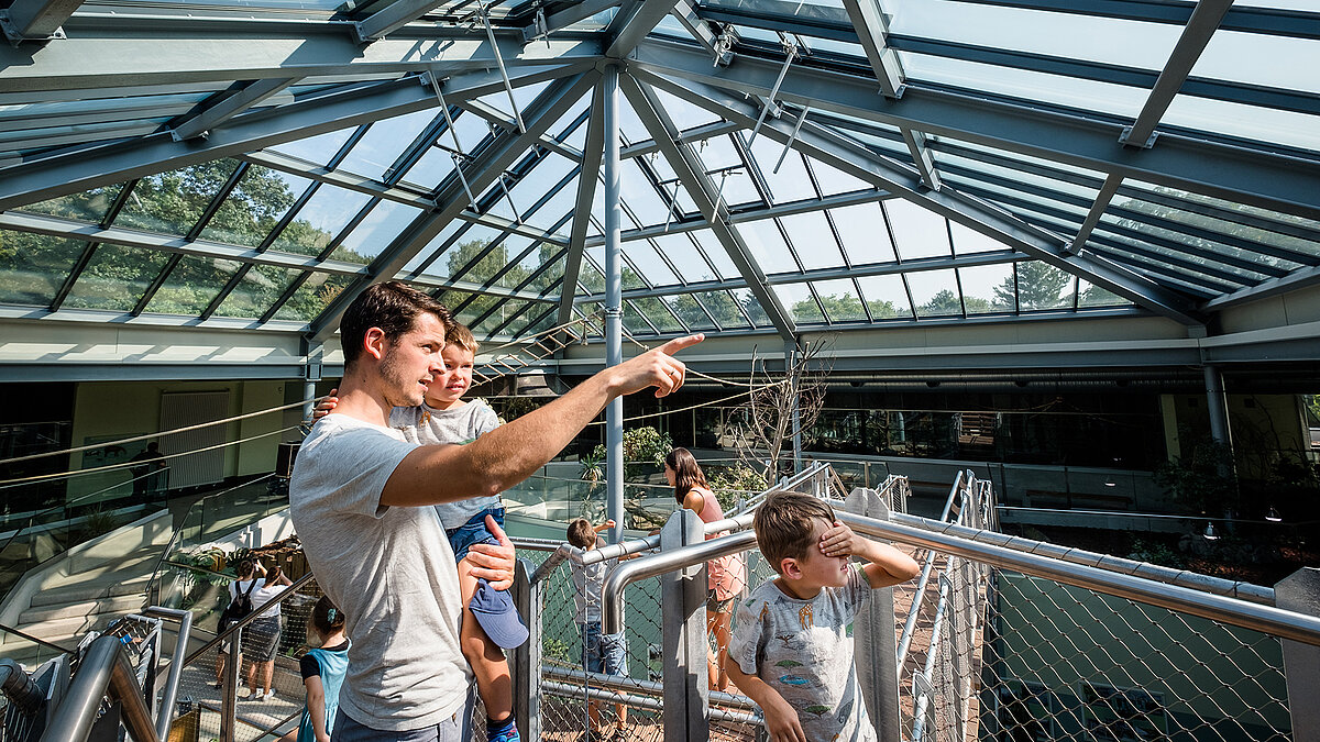 Father with children under the PR60 glass structure at Zoo Neuwied, pointing at an attraction.