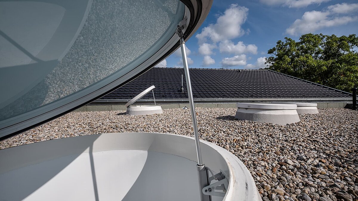 Open skylight on the gravel roof of the Sonnenfeld daycare center in Wandlitz with blue sky and white clouds.