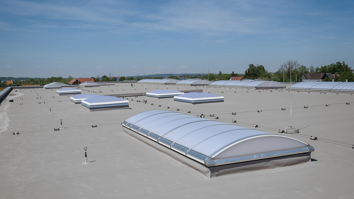 Roof of the Unglehrt construction company's production hall with multiple LAMILUX F100 W skylights and B light bands under a blue sky.
