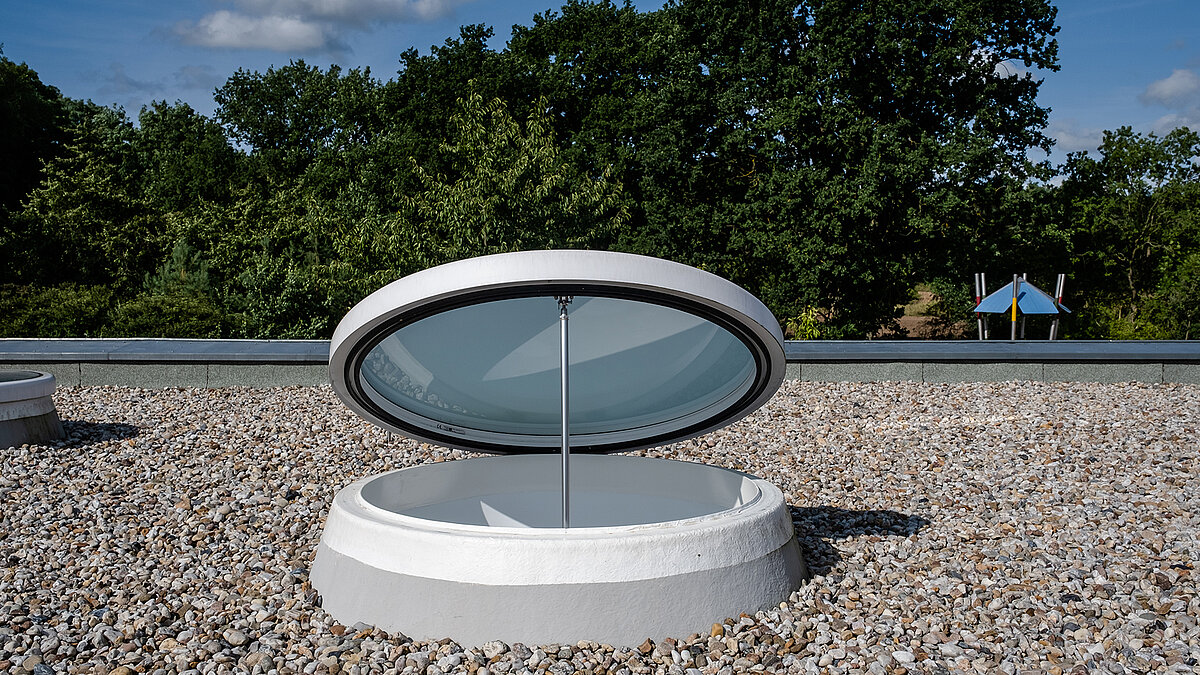 Open round skylight on the roof of the Sonnenfeld daycare center in Wandlitz, surrounded by gravel with green trees in the background.