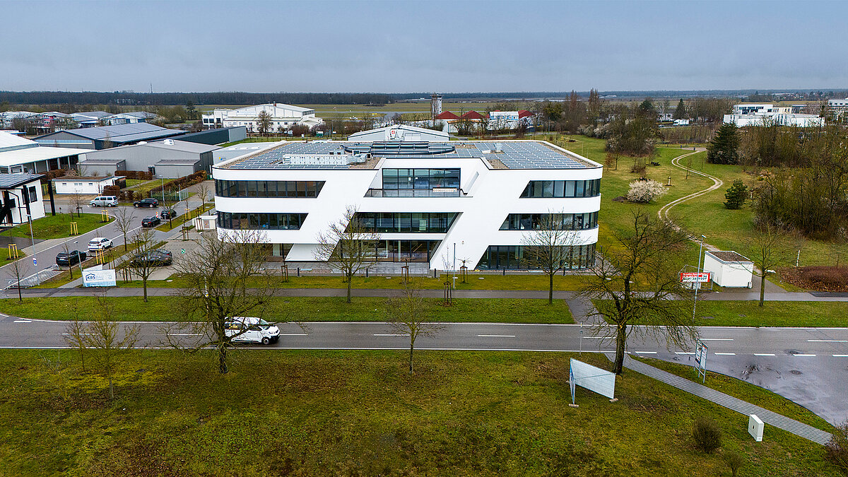 Modern office building with white facade and solar system on the roof, surrounded by green spaces and streets, under a cloudy sky.