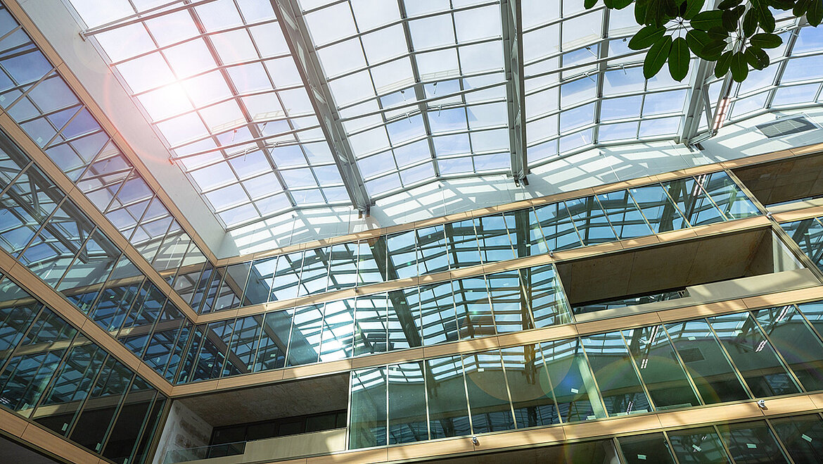 A modern building atrium with a large glass roof, multi-storey glass façades, sunlight and green plants