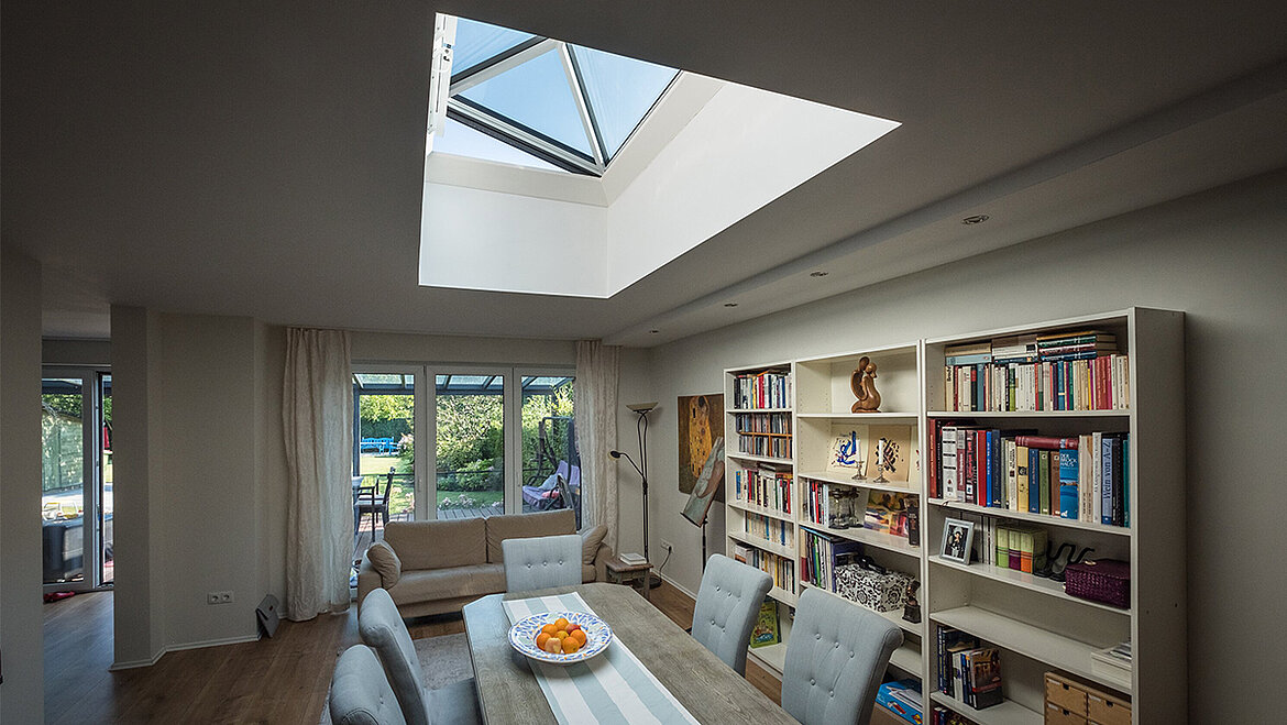 A modern living room with a skylight, a dining table and chairs, a white bookcase and panoramic windows overlooking the garden