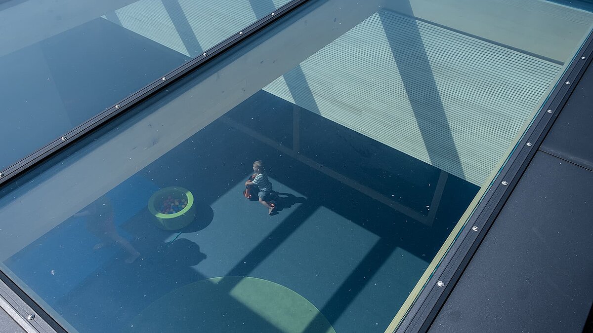 View through the large glass roof of the Wiggensbach daycare center, showing children playing and a room filled with natural light.