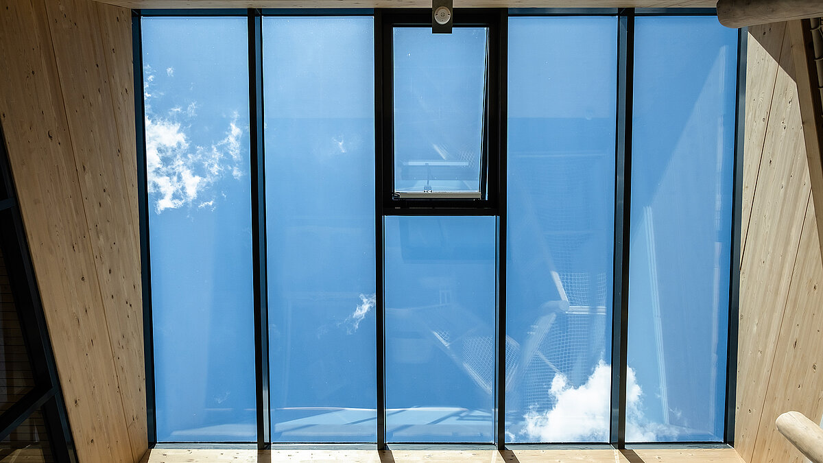 View through a large glass roof with a narrow ventilation flap, blue sky and clouds above a timber-lined interior space